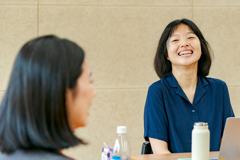 진행자 박하늘. 남색 반소매 셔츠를 입고 어깨에 살짝 닿는 단발머리를 늘어뜨렸다. 좌담 참여자를 바라보며 환하게 웃고 있다. 