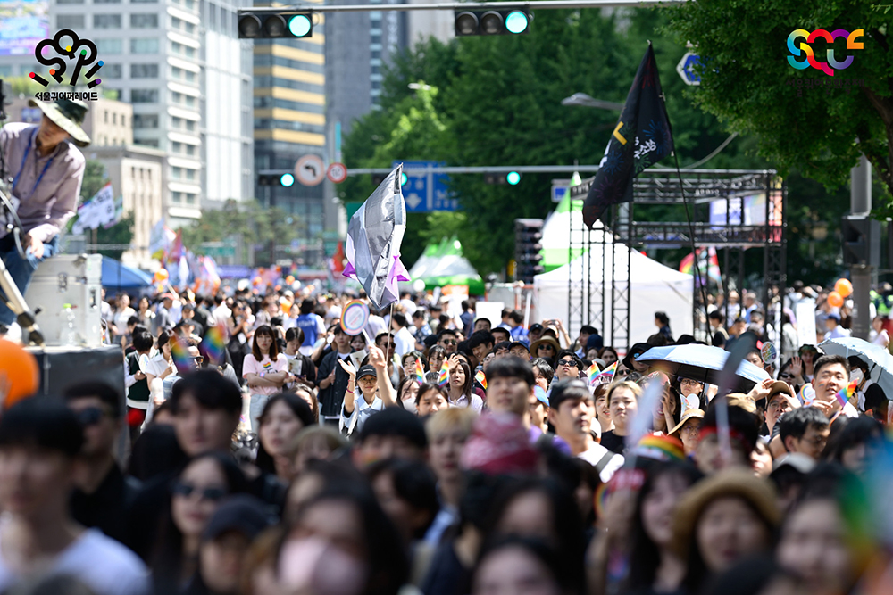 시청역에서 광화문으로 향하는 퀴어퍼레이드 행렬을 촬영한 사진이다. 사진 중앙에, 검은색, 회색, 하얀색, 보라색으로 구성된 무성애 깃발이 휘날린다. 