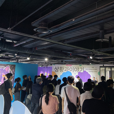 지금 들으시는 장면은, 한 전시의 개막식 현장입니다. 전시 제목은 〈상황과 이야기〉 입니다. 공간 안에는 많은 사람들이 모여 있습니다. 관람객과 관계자들은 무대처럼 보이는 현수막을 바라보며 서 있고, 현수막 앞에는 발표자와 수어를 안내하는 사람, 사회자가 서 있는 모습입니다. 관람객들은 다양한 나이대와 옷차림을 하고 있으며, 모두 조용히 앞을 주시하거나 박수를 치며 참여하고 있습니다. 무대처럼 보이는 현수막에는 다음과 같은 문구가 또렷하게 보입니다. “상황과 이야기” 서울장애예술창작센터 장애예술기획전.  참여 작가 이름이 적혀 있으며, 배경은 구름 모양으로 장식되어 있습니다. 무대 오른편에는 창문이 있어 자연광이 실내로 들어오고, 왼쪽 벽면은 푸른색 벽과 전시의 작품도 살짝 보입니다. 특히 왼편 벽에는 커다란 동물의 얼굴을 그린 회화 작품이 걸려 있어, 강렬한 눈빛이 인상적입니다. 천장에는 까만 배관과 조명이 드러나 있으며, 조명이 무대와 관람객 쪽을 은은하게 비춥니다. 