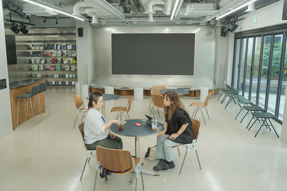 이반지하와 성수연이 원탁을 사이에 두고 서로를 마주 보고 앉아 있다. 이반지하가 오른손으로 테이블에 무언가를 그리며 말하고 있고, 성수연은 몸을 앞으로 기울여 듣는 자세다. 이들의 뒤로 커다란 검은 스크린과 서가가 위치해 있고, 곳곳에 의자와 테이블이 놓여 있다.  