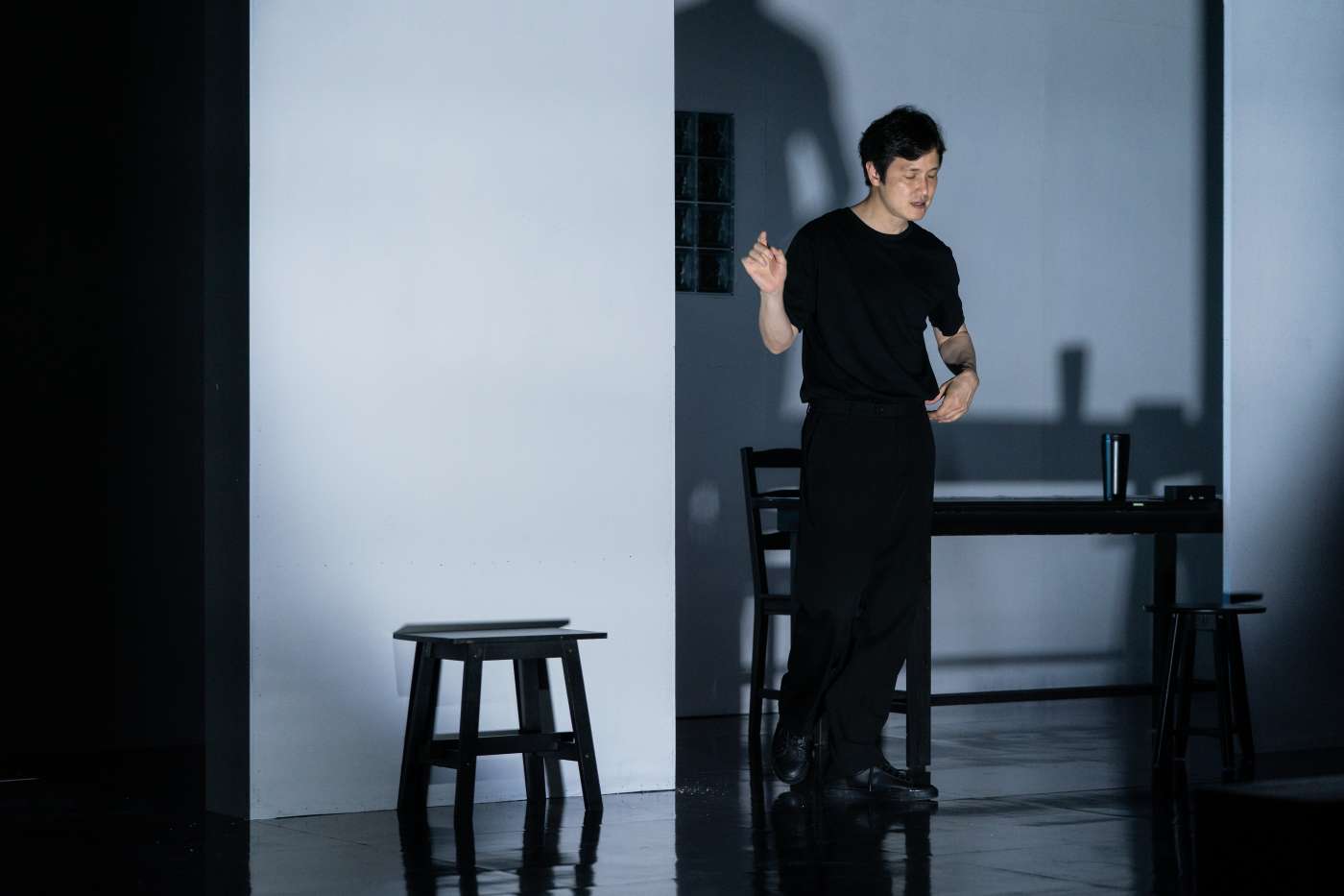 검은색 상의와 하의를 입은 남자 배우가 검은 책상과 검은 의자 앞에 서 있다. 배우는 눈을 감고 손짓하며 무언가를 말하고 있다.
