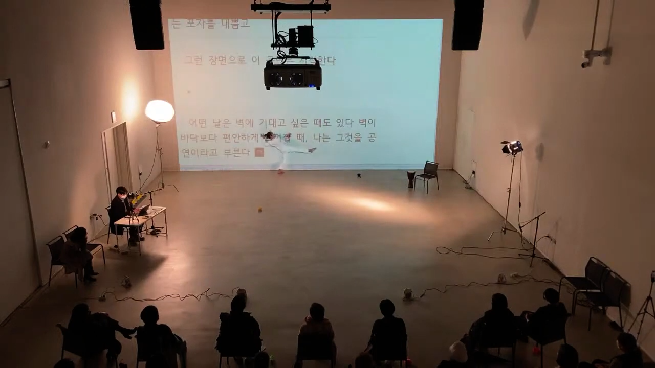 화이트 큐브 형태의 공간에서 시인과 안무가가 함께 만든 퍼포먼스가 이루어지고 있다. 정면으로 보이는 흰 벽면에는 하얀 배경 위에 검은 글씨들이 쓰이고 있다. 흰 옷을 입은 안무가는 그 글자들 앞에서 춤을 추고 있다. (또는, 그 글자들로 이루어진 벽에 기대어 있다.) 왼쪽 벽면에는 큰 조명과 테이블, 그리고 그 테이블 앞 의자에 앉은 시인이 있다. 시인은 이 이미지 아래에 인용된 시를 쓰고 있다. “어떤 날은 벽에 기대고 싶은 때도 있다 벽이 바닥보다 편안하게 느껴질 때, 나는 그것을 공연이라고 부른다” 