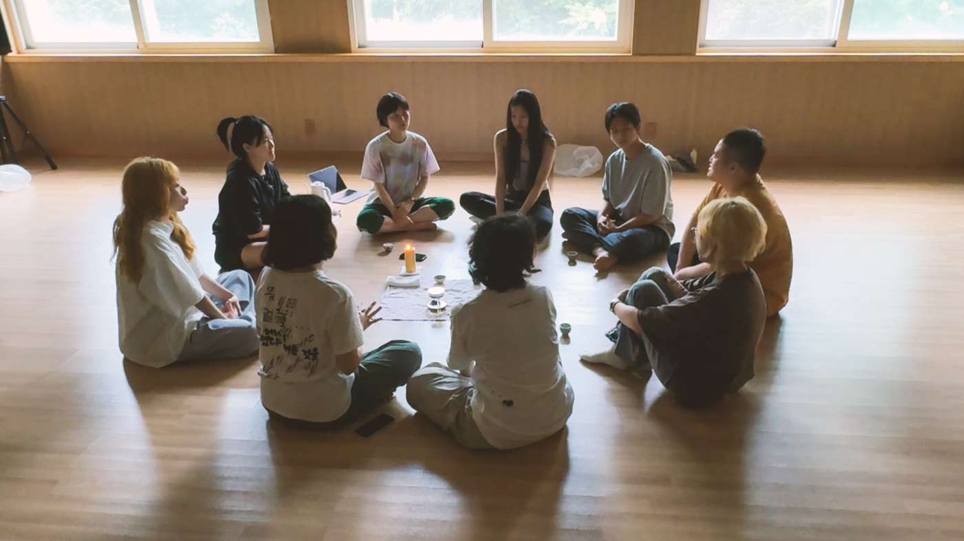 아홉 명의 사람들이 바닥에 동그랗게 모여 앉아 이야기를 나누고 있다. 그들 앞에는 작은 찻잔들과 양초가 놓여 있다.