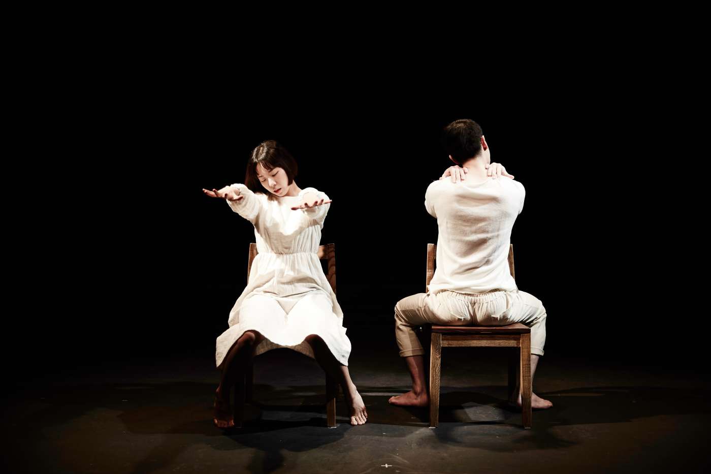 검은 무대 위, 흰옷을 입고 나무 의자에 앉은 두 배우가 있다. 단발머리의 여성 배우는 양팔을 앞으로 쭉 뻗고 무대 쪽을 바라보고 앉아 있다. 짧은 머리의 남성 배우는 양손을 어깨 위에 올린 채, 무대 뒤쪽을 바라보고 앉아 있다.