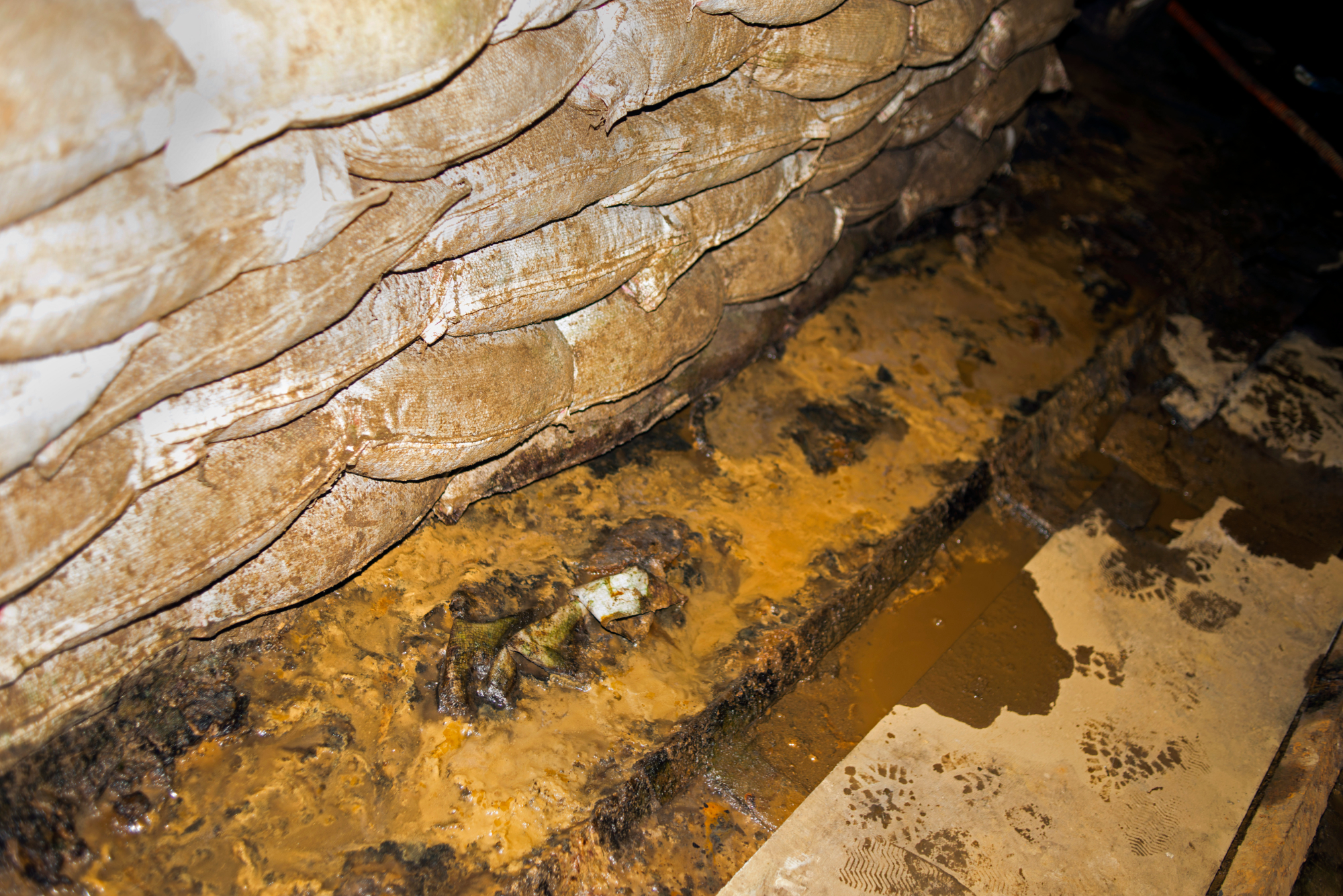 경산 코발트 광산 동굴 내부를 근거리에서 포착한 사진이다. 화면 좌측에는 미수습 유해와 토사를 수습한 밝은색 모래주머니들이 층층이 쌓여 벽을 이루고 있으며, 바닥면은 황토색 진흙과 고인 물이 뒤섞여 축축하고 생생한 질감을 가감 없이 드러낸다. 반면 우측의 건조한 지면 위로는 누군가의 행적을 암시하는 신발 자국들이 여러 개 찍혀 있다. 강한 대비 효과로 인해 현장의 거칠고 긴장감 있는 분위기를 전달한다.
