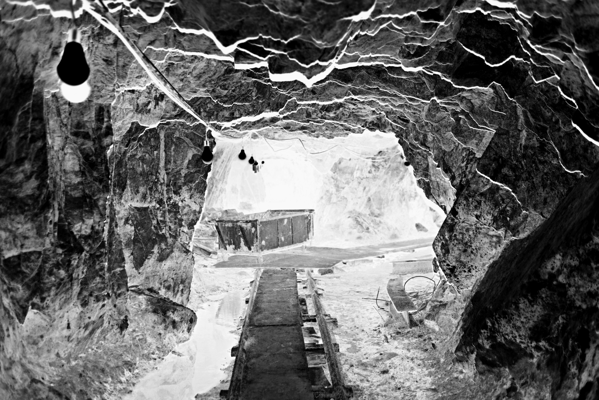 흑백 네거티브 기법으로 동굴 내부를 포착한 사진이다. 음화(네거티브) 기법의 특성상 명암이 반전되어 밝은 부분이 어둡게, 어두운 부분이 밝게 표현되었다. 이에 따라 동굴의 거친 질감과 갈라진 틈새가 더욱 날카롭게 드러난다. 바닥 중앙에는 철길 형태의 목제 발판이 동굴 안쪽으로 이어져 있으며, 그 끝에는 작업용 수레가 놓여 있다. 천장에 매달린 전구들은 네거티브 효과로 인해 검은 점처럼 묘사되어 동굴 속 빛과 어둠의 대비를 극적으로 연출한다.