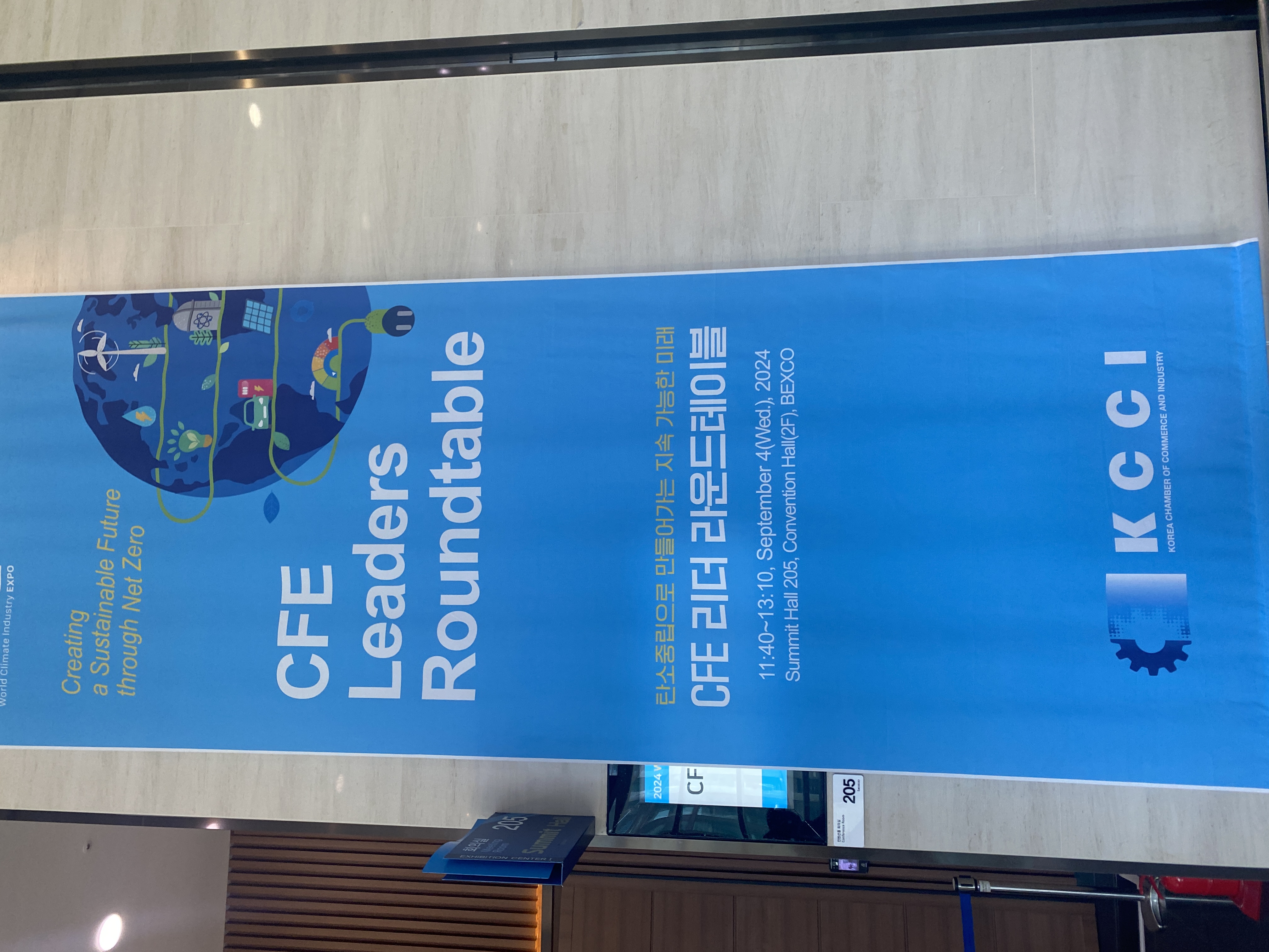 건물 내부에 걸려있는 행사 안내용 파란색 현수막. ‘CFE Leaders Roundtable’(CFE 리더 라운드테이블)이라는 행사명과 ‘탄소중립으로 만들어가는 지속 가능한 미래’라는 부제가 적혀 있다. 제목 옆 일러스트에는 초록색 콘센트와 자동차, 풍력발전소 등이 그려져 있다.