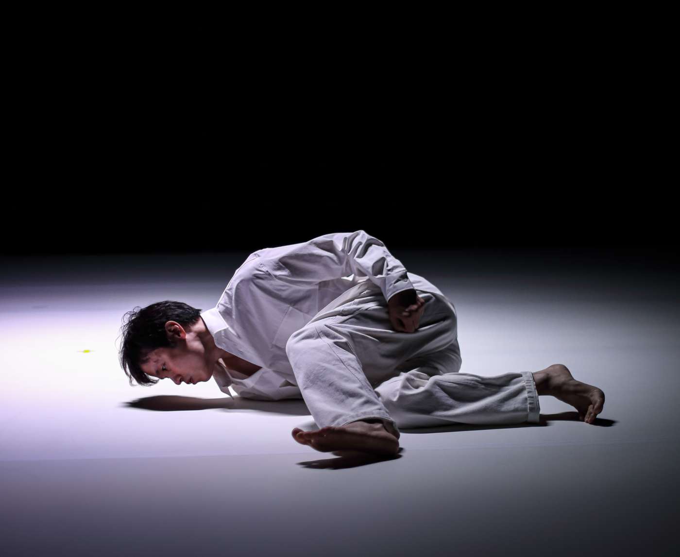 흰 바닥 위, 흰 셔츠와 흰 바지를 입은 남자 배우가 바닥에 옆으로 누워 있다. 배우는 바닥 가까이에서, 자신의 그림자를 응시하고 있다.