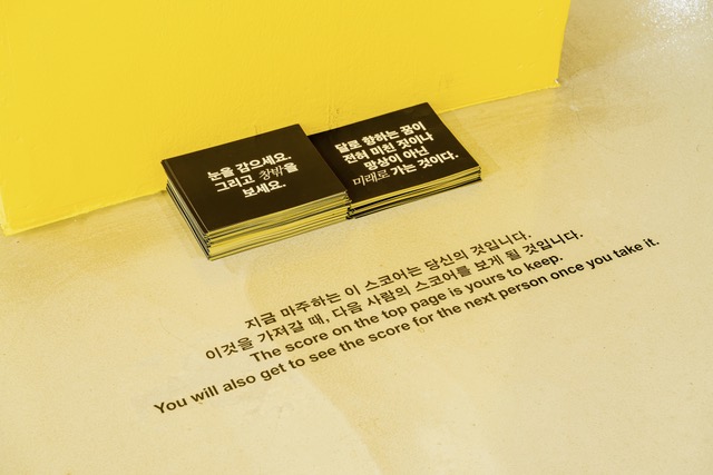 노란색 벽면 앞, 미색 바닥 위에 인쇄물 더미들이 놓여 있다. 왼쪽에 놓인 인쇄물 더미의 맨 윗장에는 검은 배경에 흰 글씨로, “눈을 감으세요. 그리고 창밖을 보세요.”라고 쓰여 있고 이중 ‘창밖’은 기울임꼴로 강조되어 있다. 오른쪽에 놓인 인쇄물 더미에는 “달로 향하는 꿈이 전혀 미친 짓이나 망상이 아닌 미래로 가는 것이다.”라고 쓰여 있고 이중 ‘미래로’는 기울임꼴로 강조되어 있다. 인쇄물들 앞 바닥에는 “지금 마주하는 이 스코어는 당신의 것입니다. 이것을 가져갈 때, 다음 사람의 스코어를 보게 될 것입니다.”라는 말이 국문과 영문으로 적혀 있다. 