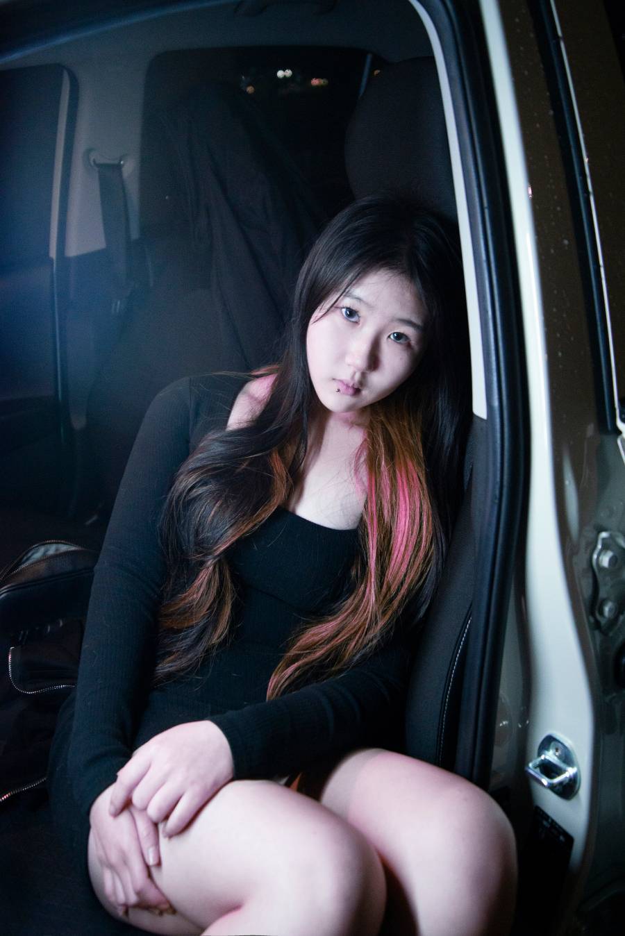 자동차 안, 검은 옷을 입고 긴 머리 카락의 일부를 붉게 염색한 사람이 앉아 있다. 입술 아래에는 피어싱을 했다.
